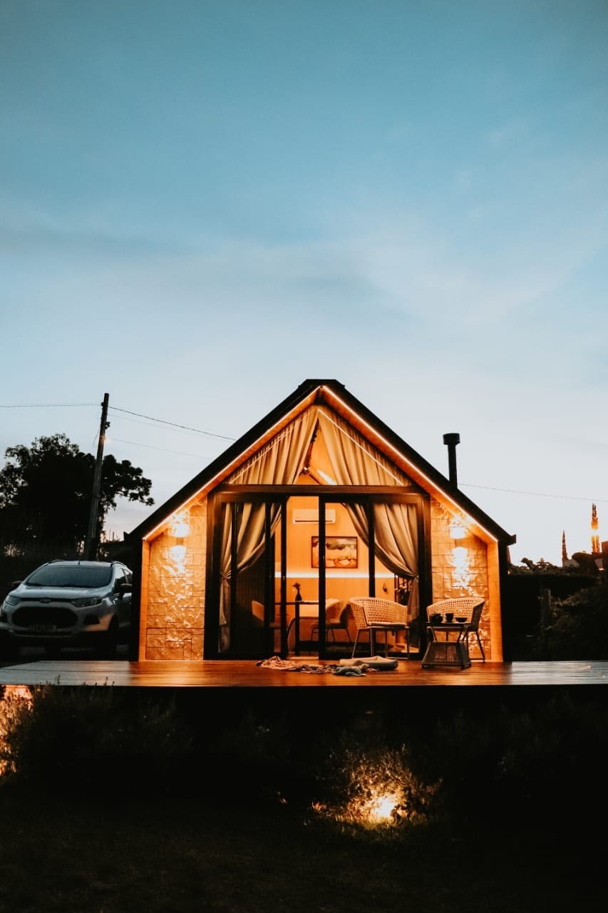 A charming cabin is bathed in warm light as twilight sets in. A spacious deck features two comfortable chairs positioned at the entrance, inviting relaxation. The large glass doors provide a glimpse into a cozy interior, framed by soft curtains that flow gently in the evening breeze.