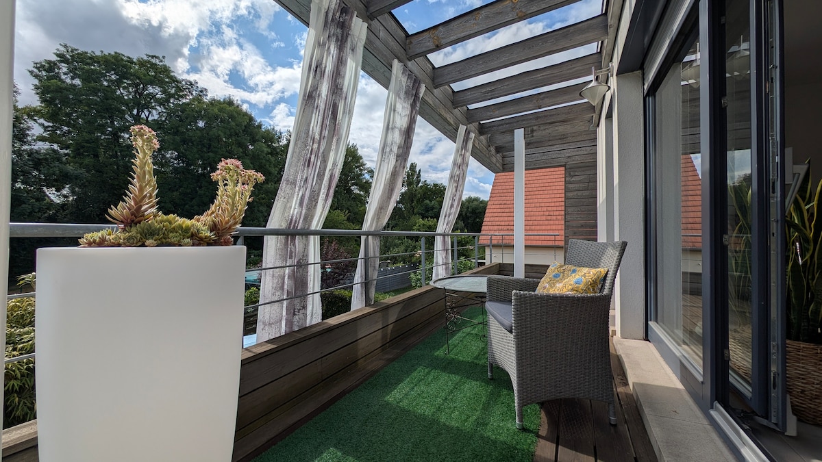 A terrace is shown with modern wooden beams and a railing, featuring a cozy seating area with a patterned cushion. Potted plants add greenery, while a view of trees and rooftops is visible in the background under a partly cloudy sky.