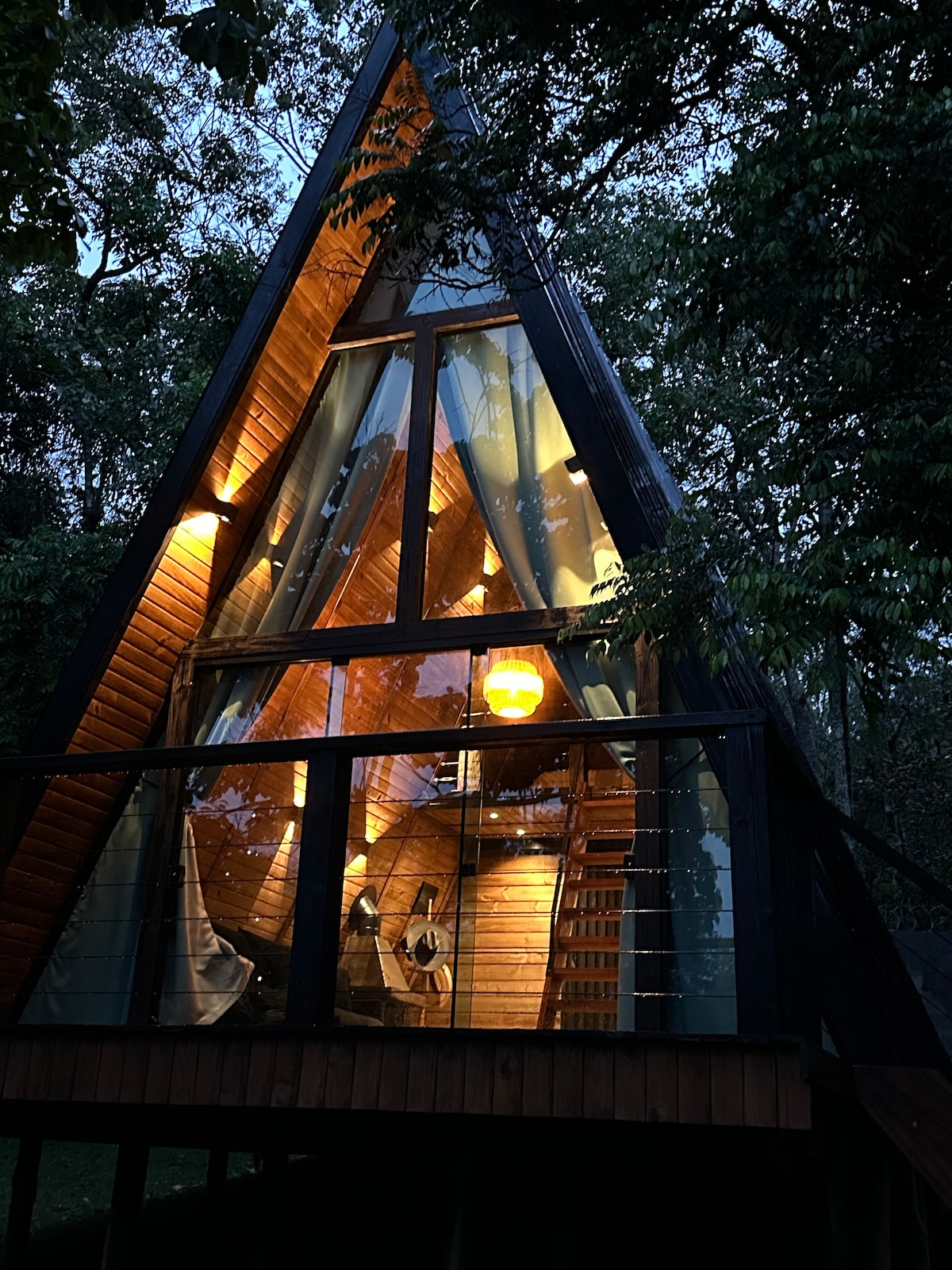 A triangular wooden cabin is surrounded by trees, creating a secluded atmosphere. Warm light glows from within, illuminating the interior features through large glass panels. A balcony extends from the structure, offering views of the natural surroundings.