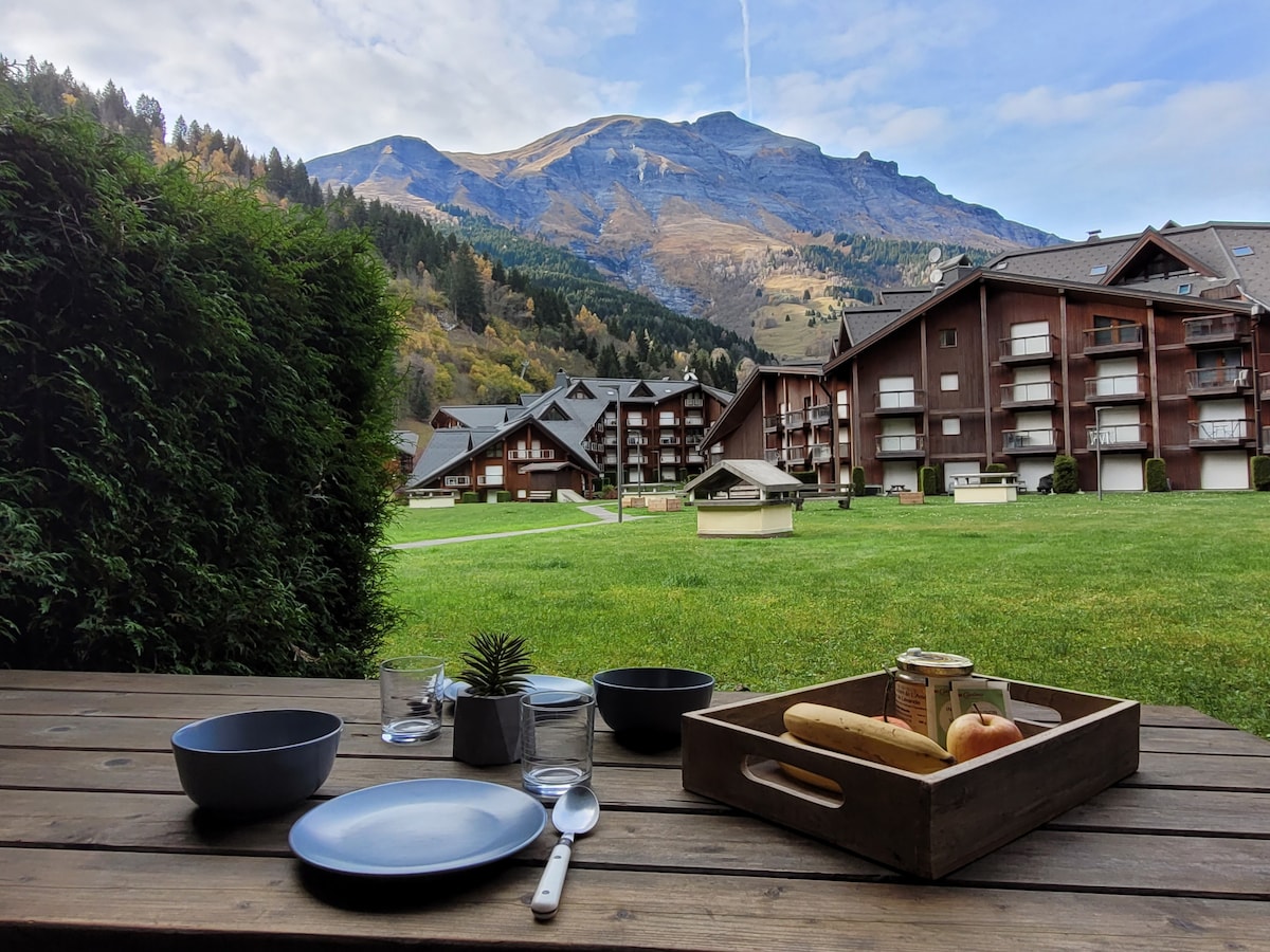 A wooden terrace table is set with plates, cutlery, and a snack tray. The background features a scenic view of mountains and well-maintained green lawns. Several chalet-style buildings are visible, blending harmoniously with the natural landscape.