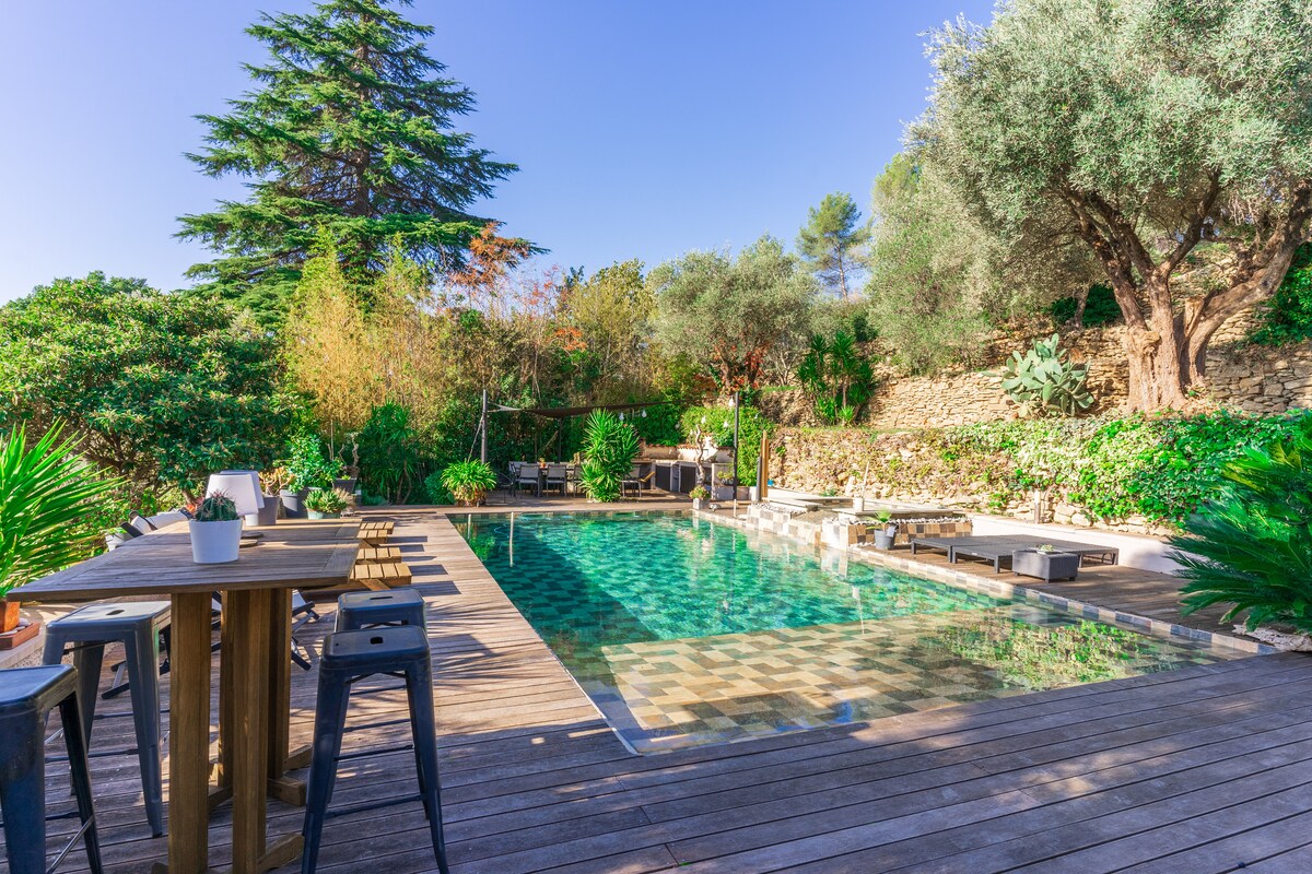 An outdoor pool is surrounded by wooden decking and lush greenery. Several lounge chairs are positioned near the pool, and a dining table with high stools is situated nearby. The serene landscape is complemented by various plants and trees, creating a calming atmosphere.