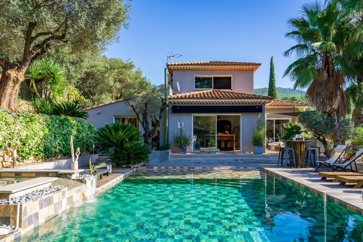 A villa is surrounded by lush greenery, featuring a tranquil swimming pool with mosaic tiles reflecting shades of blue and green. Sun loungers and a small dining area are positioned nearby, with the structure showcasing a two-story design and traditional roof tiles.