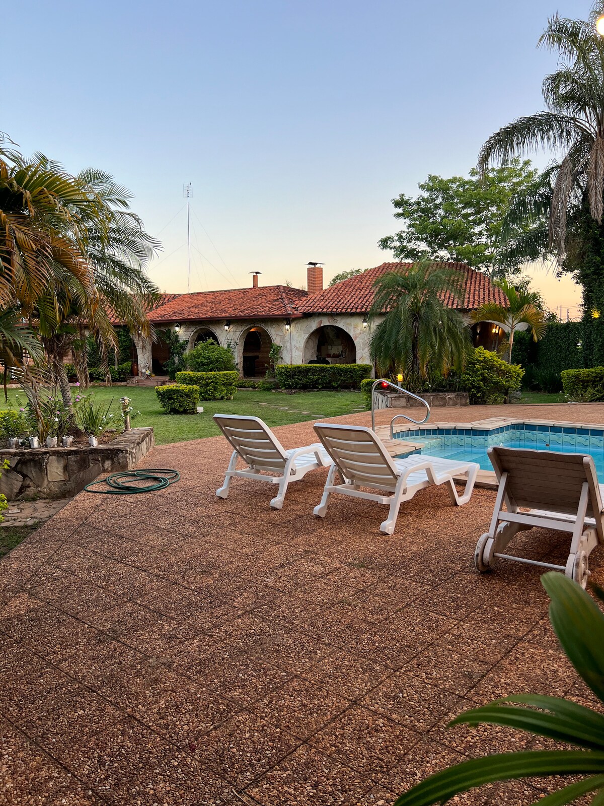 A tranquil pool area is framed by lush greenery and palm trees. Two white lounge chairs are positioned beside the pool, inviting relaxation. The colonial-style house with a red-tiled roof is visible in the background, surrounded by manicured gardens.