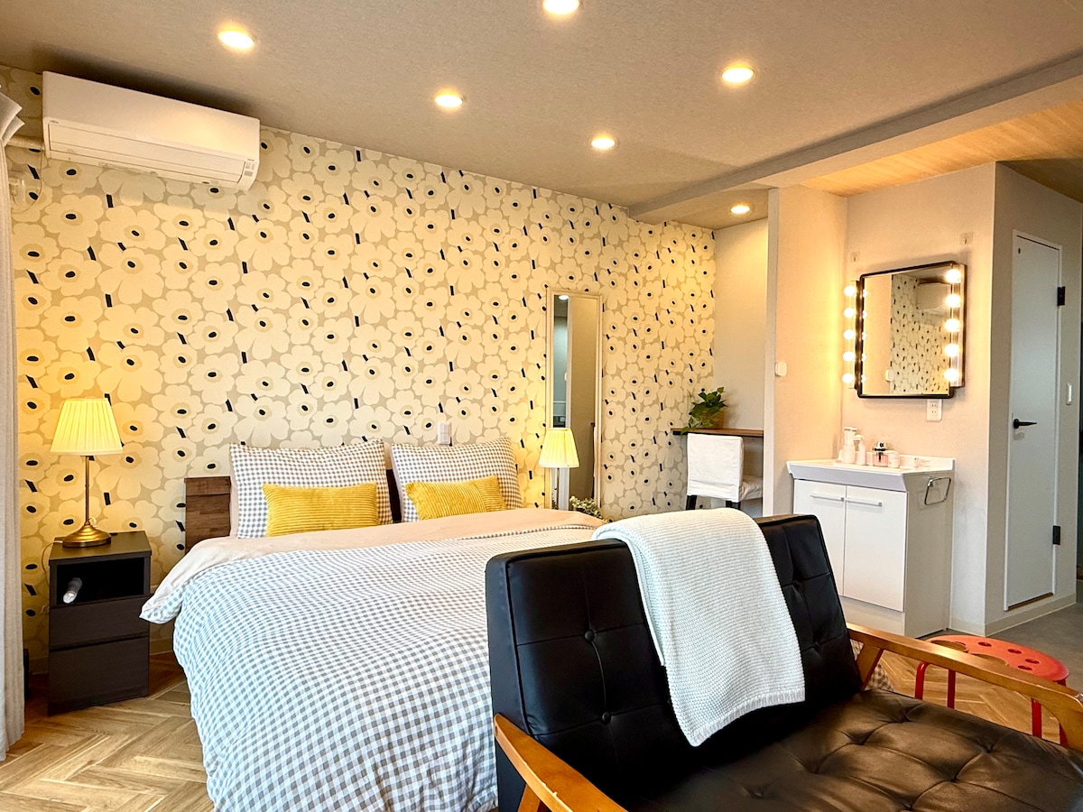 A comfortable sleeping area features a queen-size bed with a checkered bedspread and two decorative pillows. A brightly patterned wall and mirrored closet add visual interest. The space includes a cozy double sofa and a well-lit vanity area, providing a functional and inviting atmosphere.