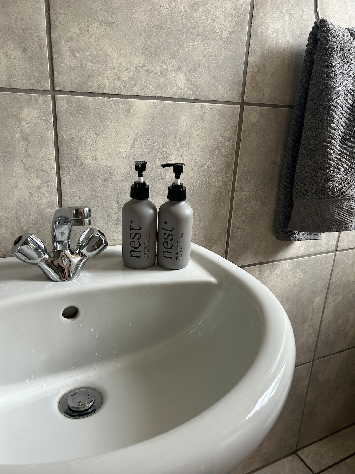 A clean sink is positioned against tiled walls, featuring a chrome faucet and two gray bottles of body wash and shampoo labeled 'nest.' A textured gray towel is neatly hung beside the sink, contributing to the bathroom's functional design.