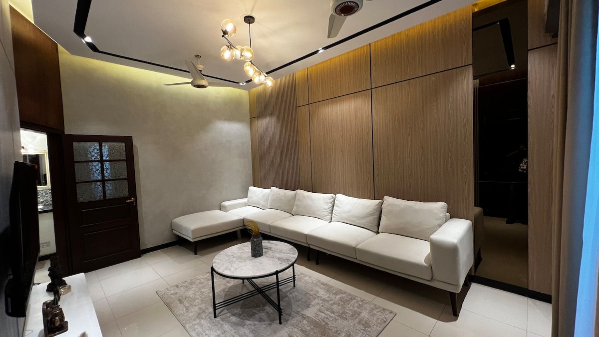 A spacious lounge area is furnished with a large white sectional sofa and a circular area rug. A modern coffee table stands at the center, while a stylish light fixture illuminates the room. Accent walls of warm wood create a calming ambiance.