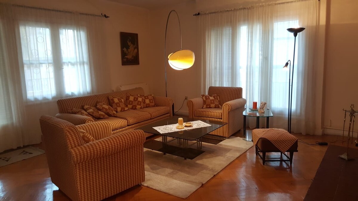 A well-furnished living area features a patterned sofa, armchair, and a coffee table. Natural light filters through sheer curtains covering two windows. A slender floor lamp stands beside the sofa, while a small side table holds decorative items.