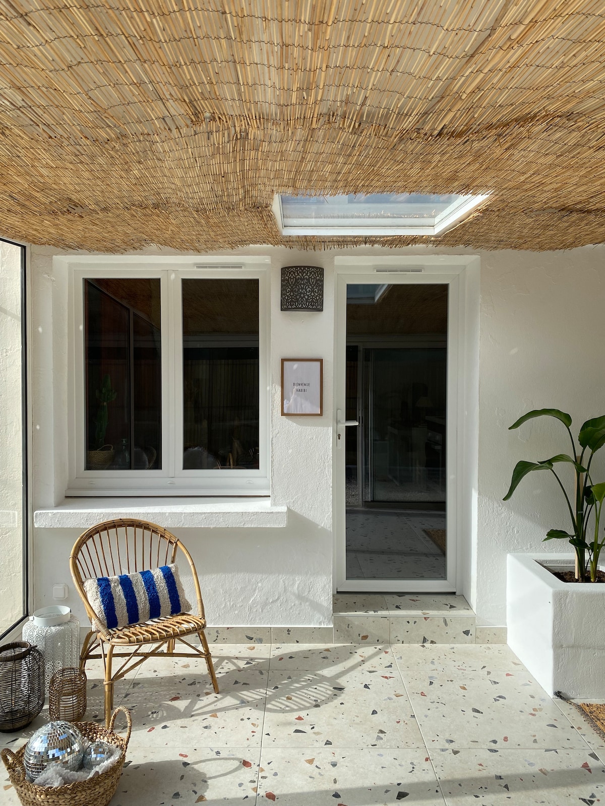 A serene entrance area features a light-filled space with a woven ceiling overhead. A rattan chair is positioned beside a potted plant, and natural light streams through large windows. Decor elements include a lantern and small decorative items, creating a welcoming ambiance.