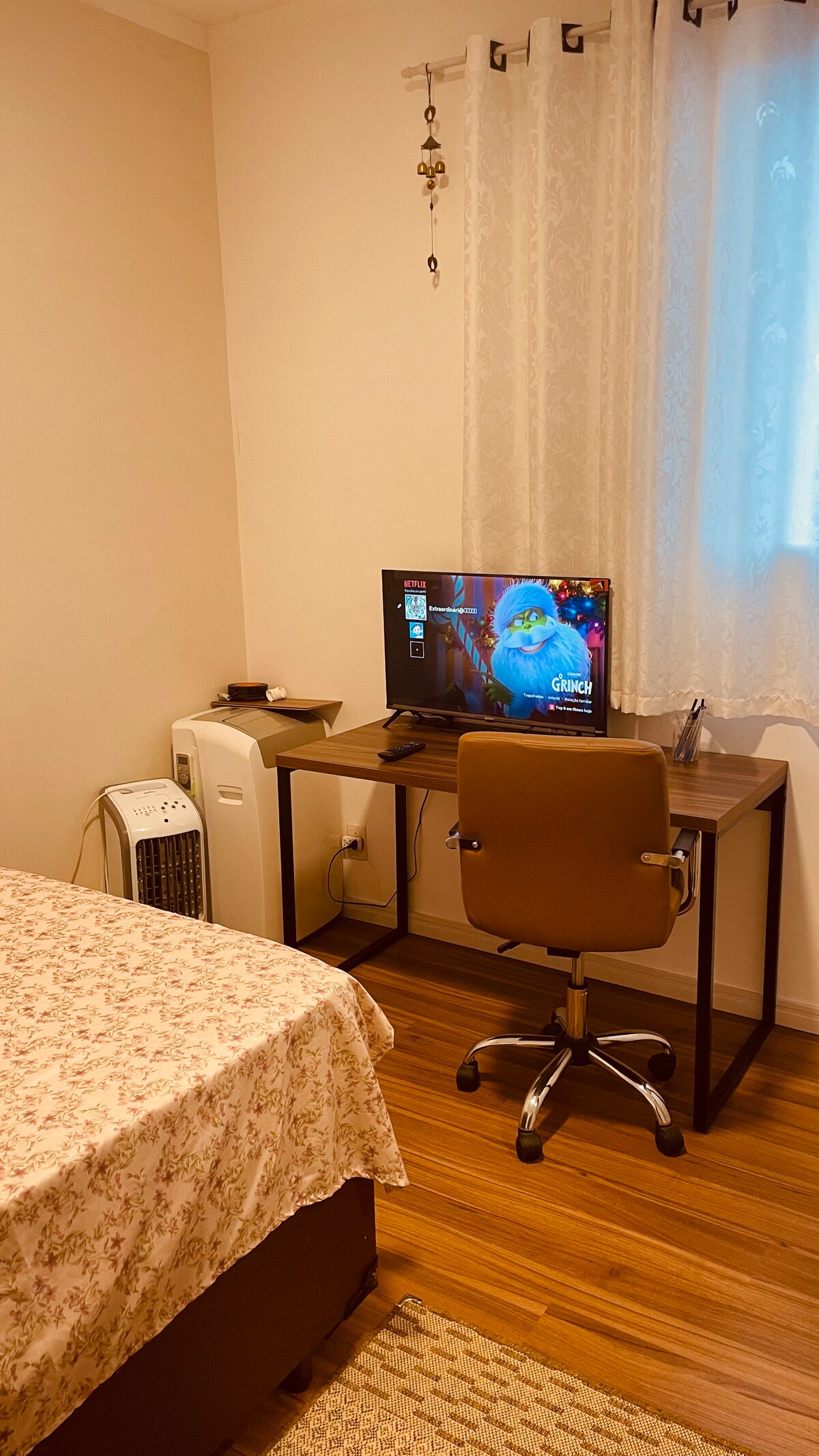 A workspace is arranged near the bed, featuring a sleek desk with a computer monitor. A comfortable chair is positioned in front of the desk. A portable air conditioning unit rests on the floor, and natural light filters through the sheer lace curtains.