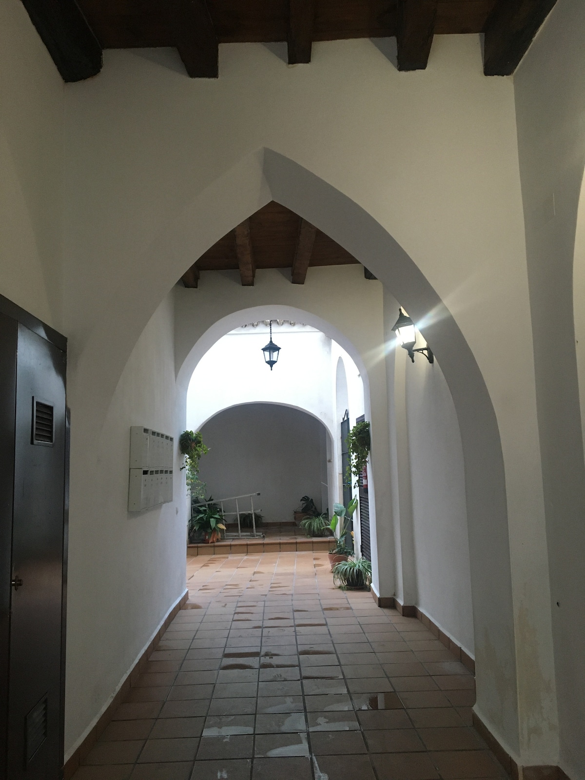 A narrow hallway features arched doorways and wooden beams overhead. Soft lighting is provided by wall-mounted lamps, and potted plants are positioned along the walls. The tiled floor reflects light, enhancing the spacious feel of the corridor leading to an open area.