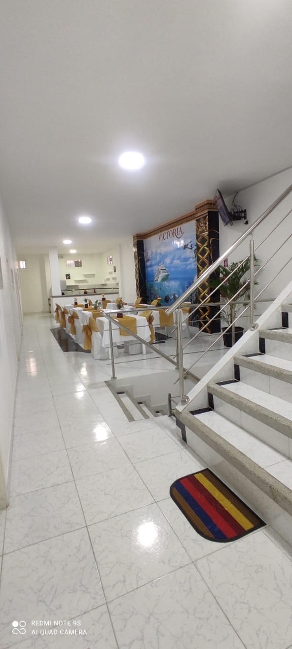 A well-lit hallway leads to an open dining area featuring neatly set tables adorned with beige and gold tablecloths. A large wall mural depicting a tropical scene adds a touch of color, while a polished staircase is visible on the right side.