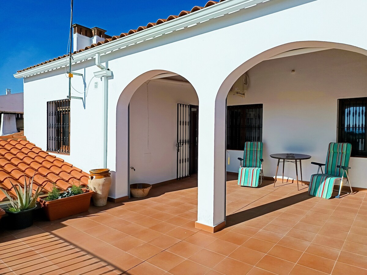 A sunlit terrace is presented, featuring two striped lounge chairs and a small round table. The space is framed by white arches and terracotta roofing, with potted plants adding greenery. A clear blue sky creates a bright, open atmosphere.