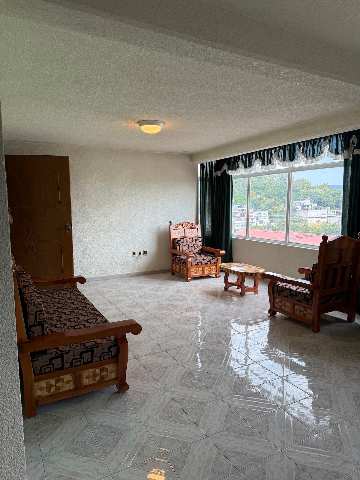 A small sitting area is furnished with two wooden chairs and a compact table, positioned near a large window. Natural light filters in, illuminating the room's glossy tiled floor and simple decor.