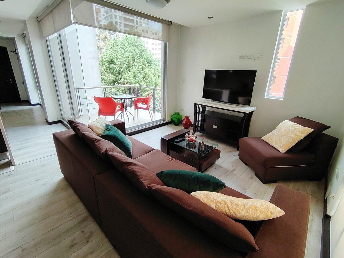 A spacious living area includes a comfortable brown sofa adorned with colorful cushions. A modern coffee table sits at the center, complemented by a green plant. Natural light enters through large windows, and a balcony is visible, furnished with red chairs.