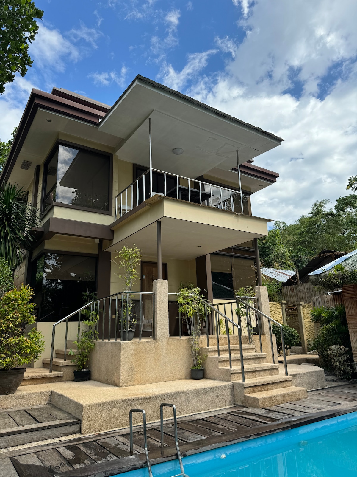 The spacious beach home is displayed with a modern architectural design. A short set of stairs leads to the entrance, surrounded by greenery. Large windows provide natural light, and a clear pool is visible in the foreground, reflecting the blue sky and clouds.