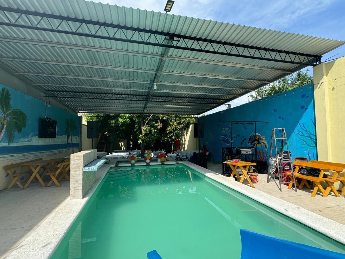 A covered pool area features a clear water pool with seating along the sides. Two yellow chairs are positioned beside the pool, and several wooden picnic tables are arranged nearby. Colorful murals depicting underwater scenes adorn the walls.