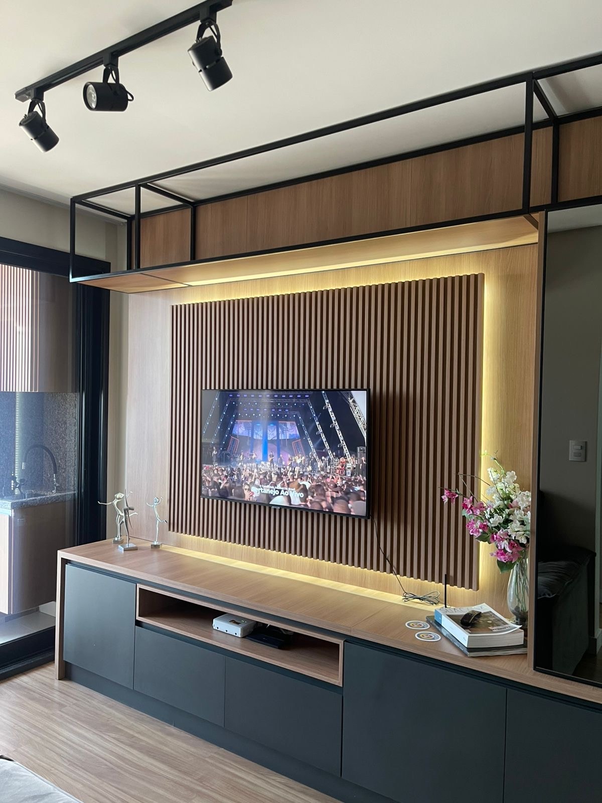 A modern living area features a wall-mounted television set against a textured wooden backdrop. Soft lighting highlights the entertainment unit below, which includes decorative items and a plant, adding a touch of elegance to the space.
