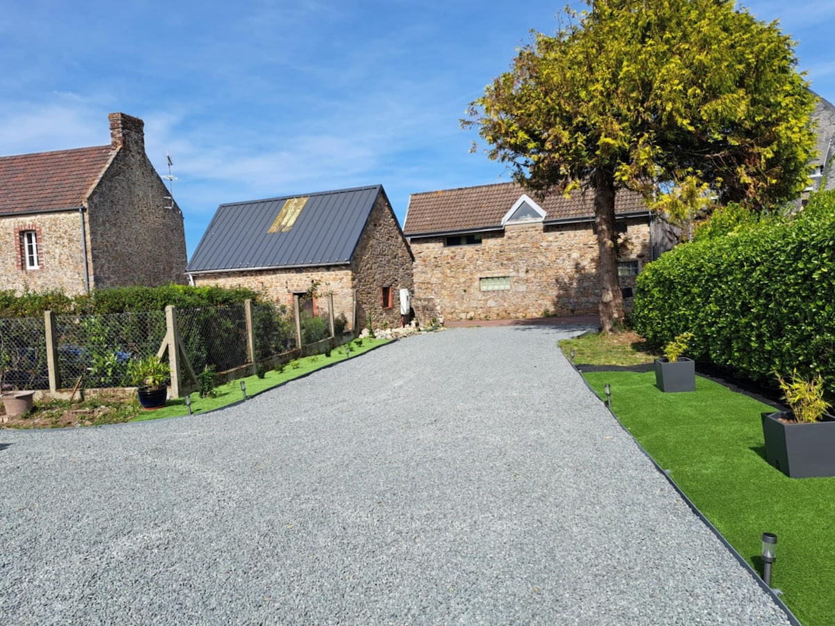 A wide gravel pathway leads through a neat landscape, flanked by greenery and small planters. Stone buildings with varied rooftops provide a rustic backdrop, accented by a large tree casting shade on the right. The clear sky enhances the inviting outdoor space.