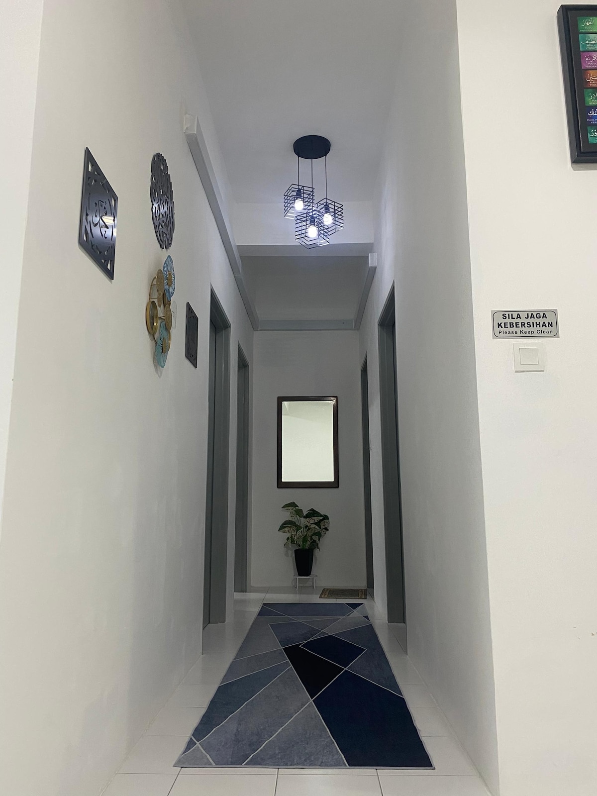A well-lit corridor features a modern ceiling light and a decorative rug with geometric patterns. Framed artwork adorns the walls, and a mirror is positioned above a small plant near the end of the hallway, enhancing the open feel of the space.