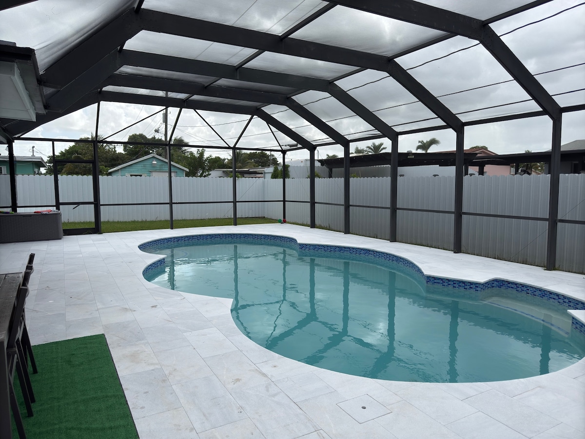A screened-in outdoor pool area is presented, featuring a uniquely shaped pool with a blue tiled edge. Surrounding the pool, tiled flooring provides a clean and spacious feel. A dining area with dark furniture is visible in the background, offering additional relaxation space.