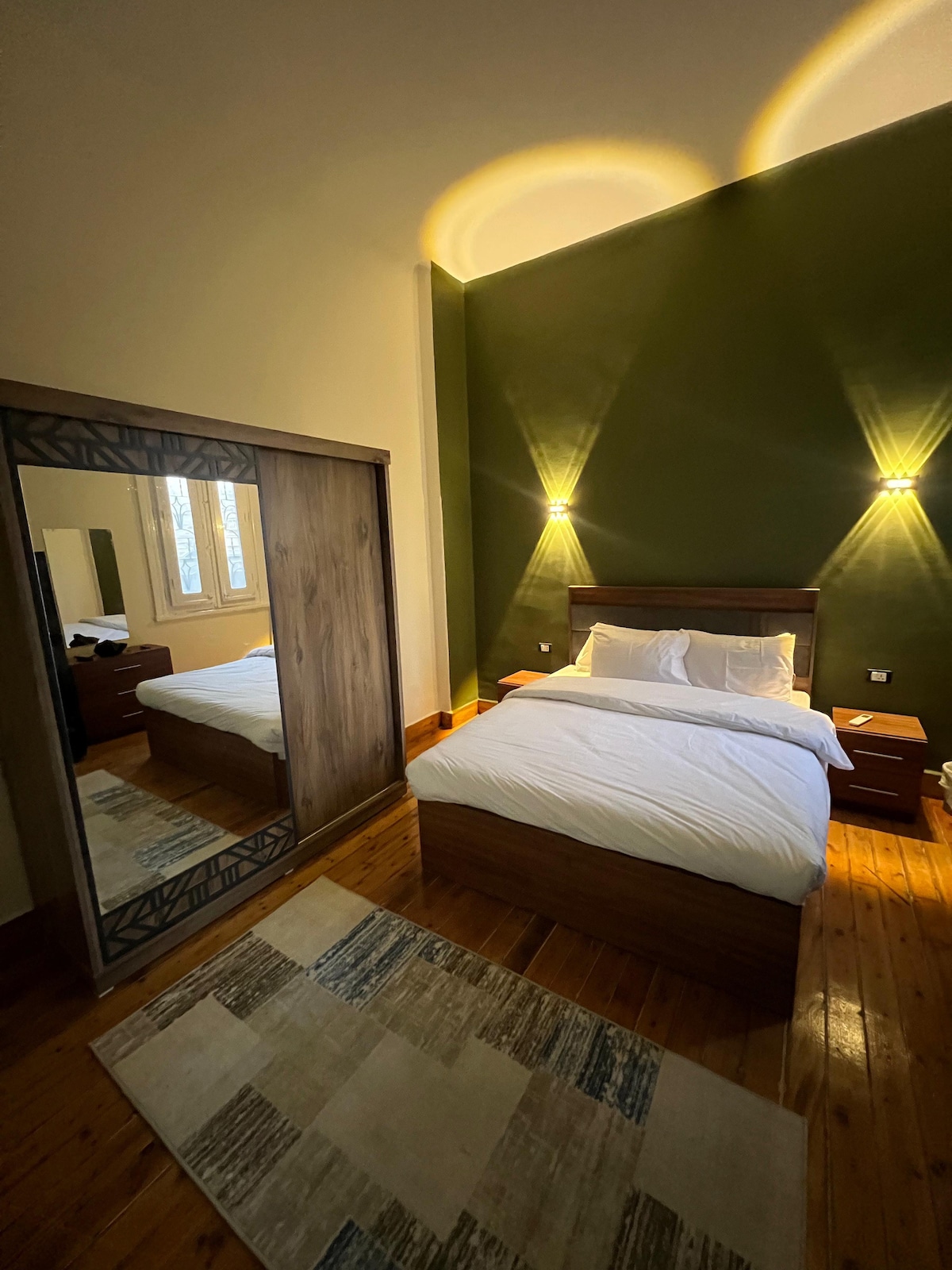 A cozy bedroom is presented, featuring a spacious bed with crisp white linens. Soft lighting is provided by wall-mounted sconces. A large mirror reflects the room, which is complemented by warm wood accents and a light-toned rug on the hardwood floor.