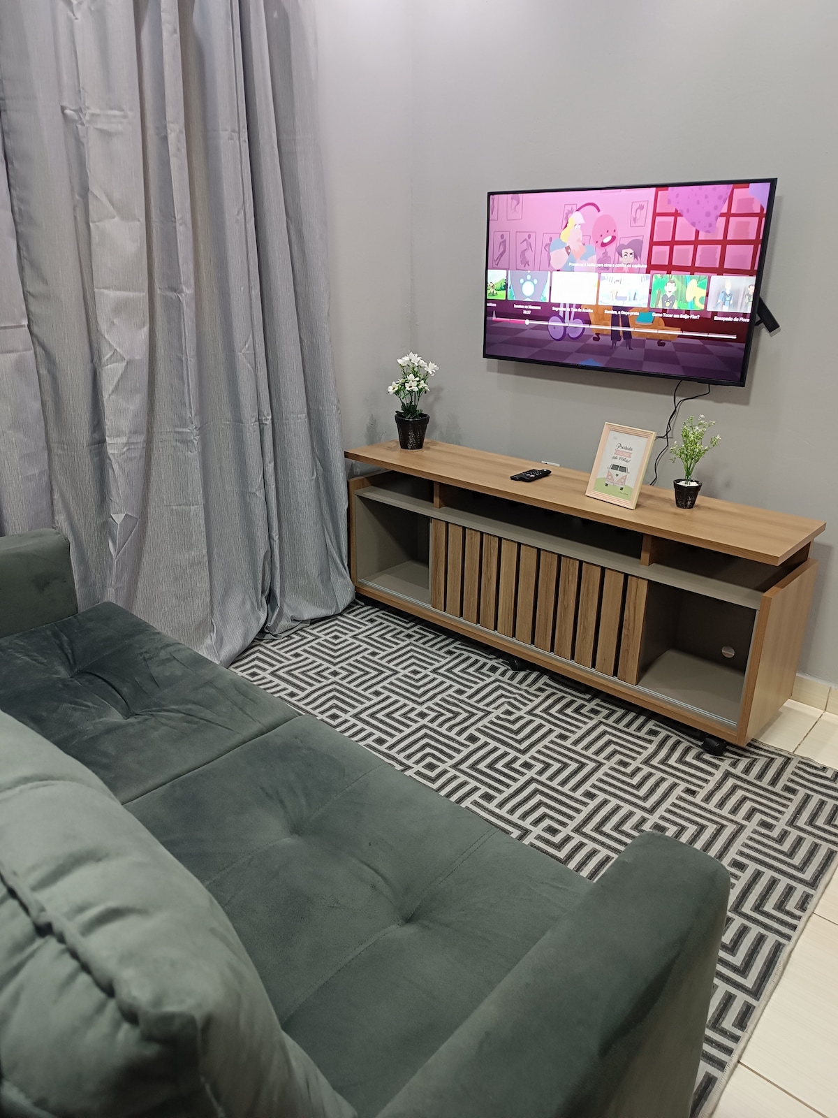 A cozy living area includes a soft green sofa facing a mounted TV, which displays colorful content. A wooden entertainment unit below holds decorative plants and a framed piece. Light gray curtains enhance the space, and a patterned rug is laid beneath.