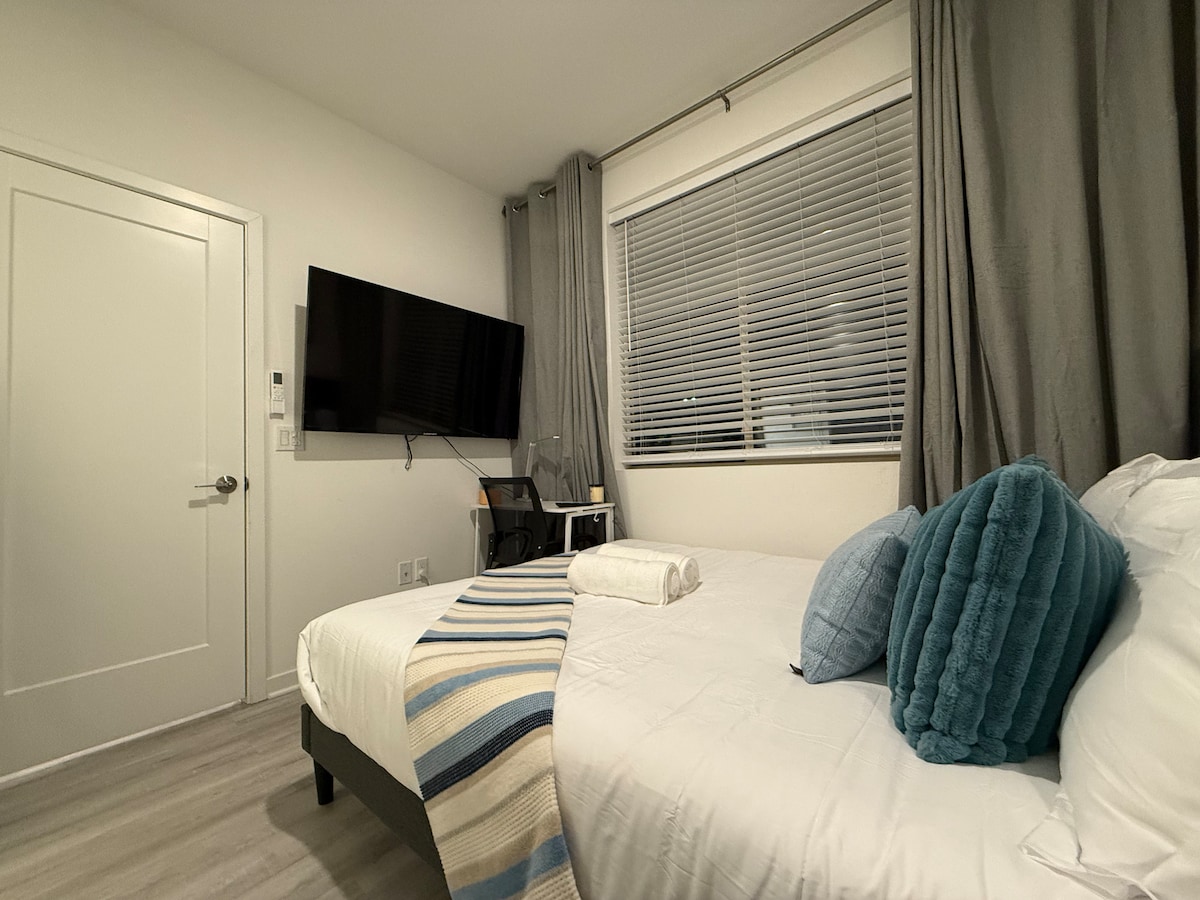 A cozy sleeping area is presented with a spacious bed topped with soft pillows and a neatly rolled towel. A 65-inch flat-screen TV is mounted on the wall, and natural light enters through a window with sheer blinds, complemented by elegant grey curtains.