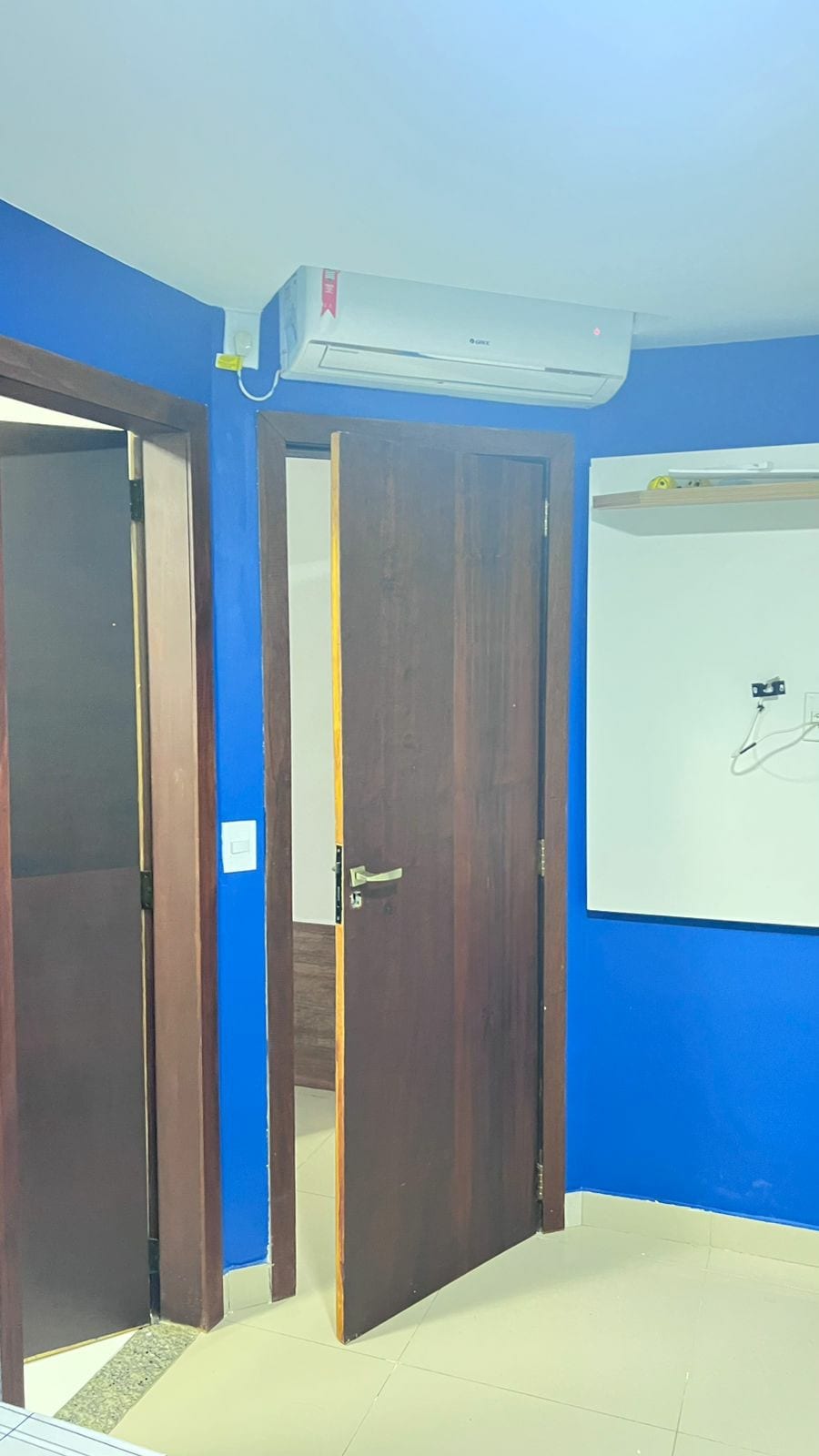 A bright blue wall is present in this room, creating a vibrant atmosphere. A wooden door leads to another space, while an air conditioning unit is mounted above. A shelf is attached to the wall, providing additional storage or display options.