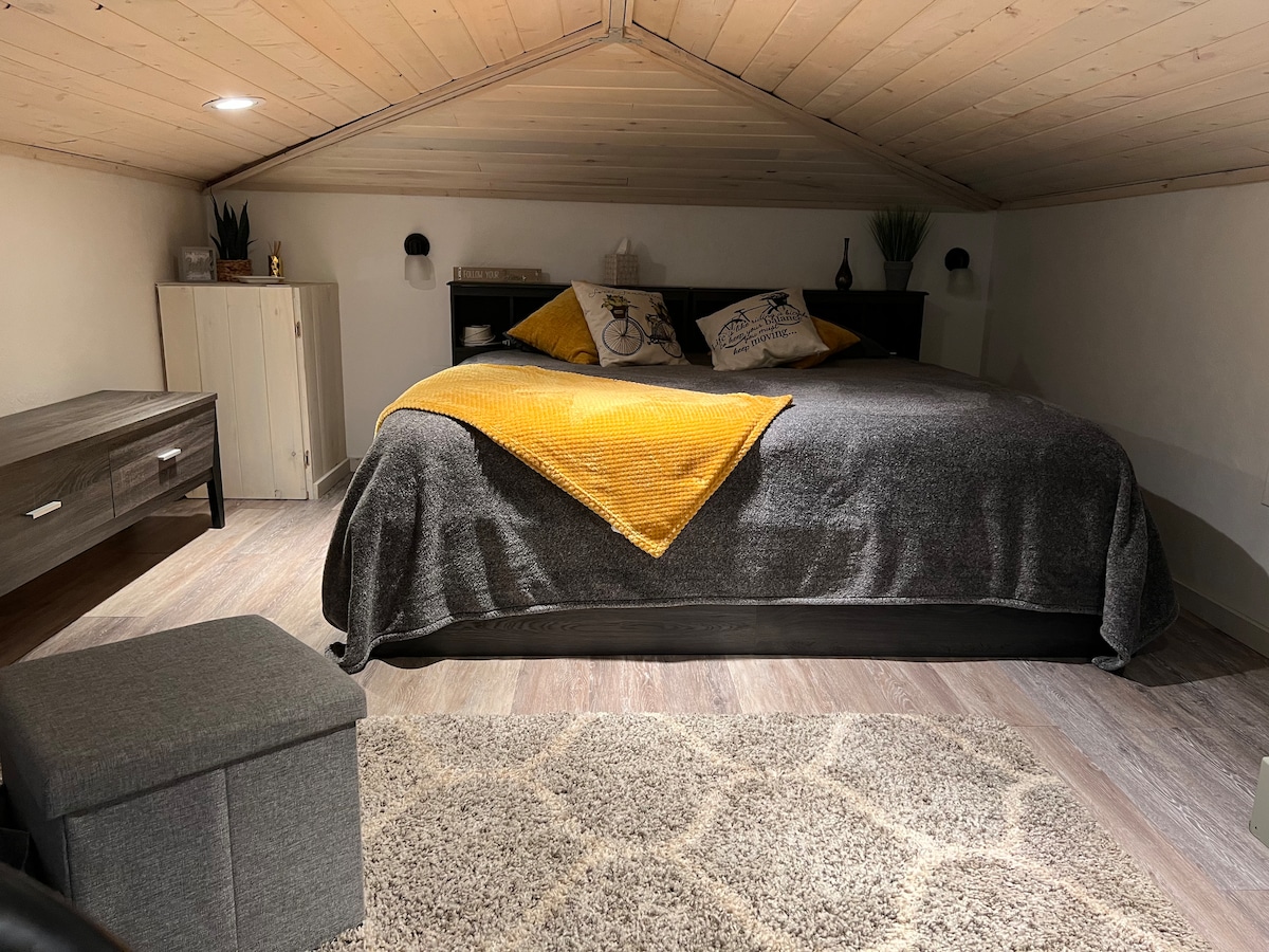 A cozy sleeping area is revealed under a sloped wood ceiling. A spacious bed is dressed in a soft gray blanket and accented with a vibrant yellow throw. A bedside table and a small storage unit are positioned nearby, contributing to the functional layout.