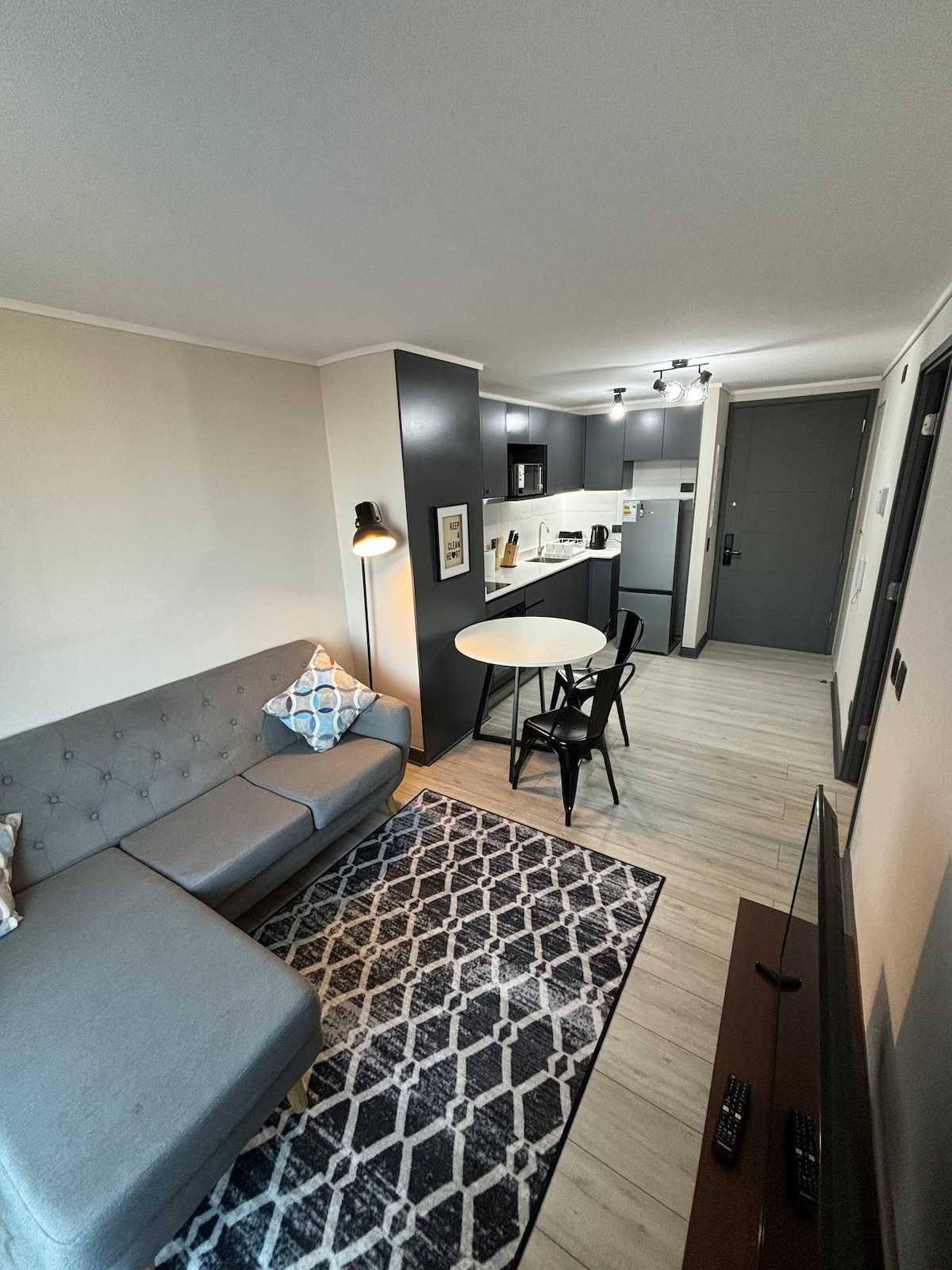A modern living space is depicted, featuring a cozy gray sectional sofa adorned with a decorative pillow. A round dining table with black chairs occupies the center, complemented by a patterned area rug. An open-concept kitchen with modern appliances is visible in the background.