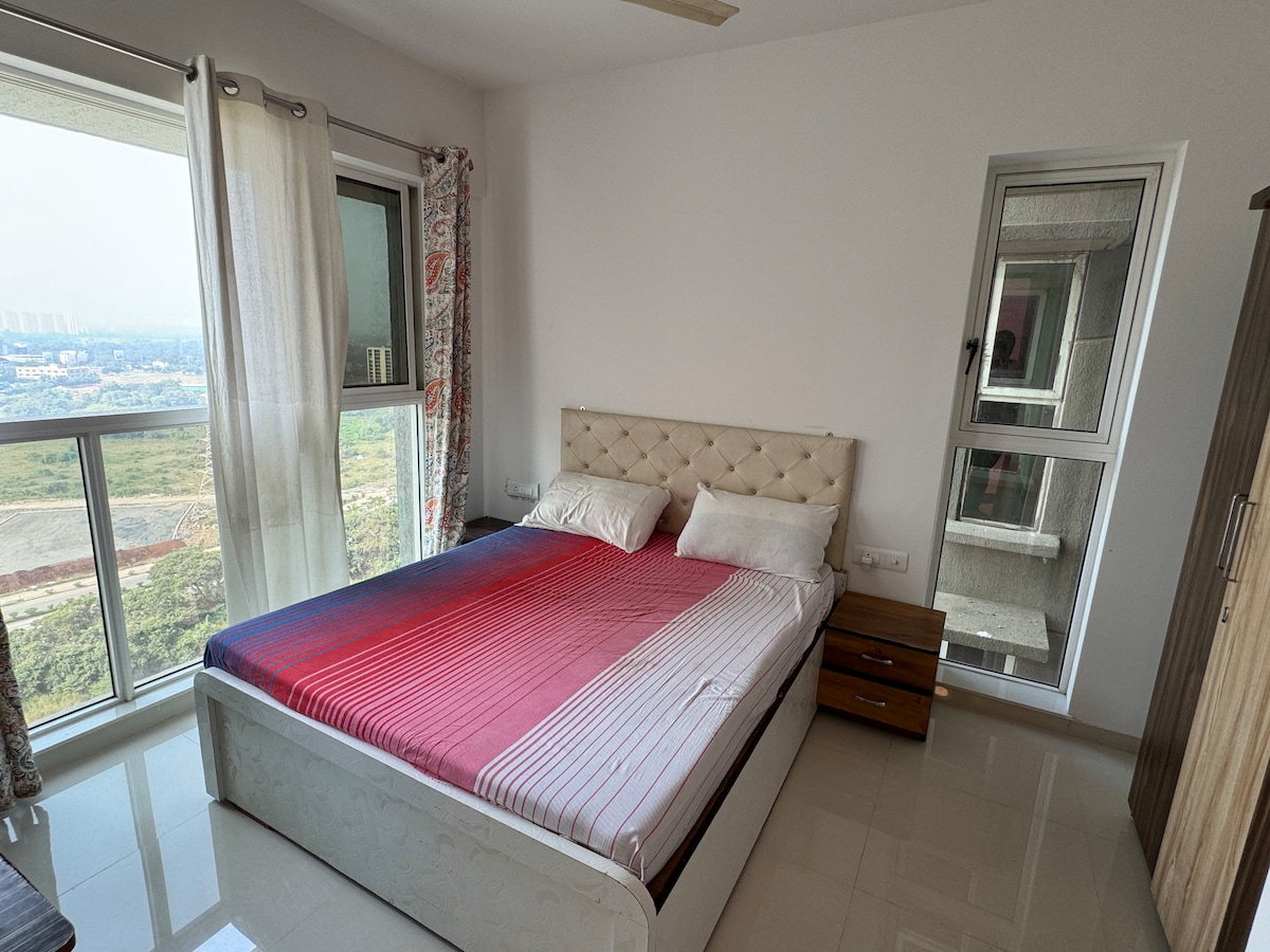 A bright bedroom is highlighted by a spacious bed with a patterned red and white bedspread. Large windows provide a view of the surrounding landscape, with sheer curtains allowing soft light to filter in. A wooden nightstand and a mirror add to the room's functionality.