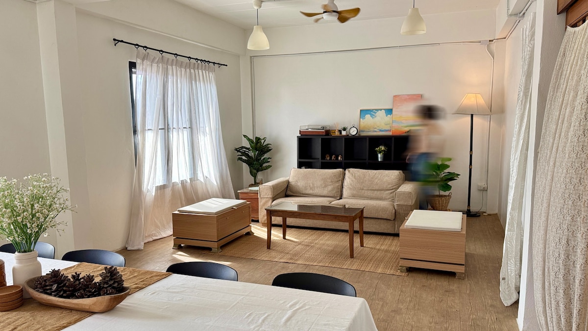 The living area features a beige sofa paired with a wooden coffee table, enhancing the space's openness. Large windows with sheer curtains allow natural light, while decorative plants add a touch of greenery. A dining area is visible, accommodating several black chairs around a table.