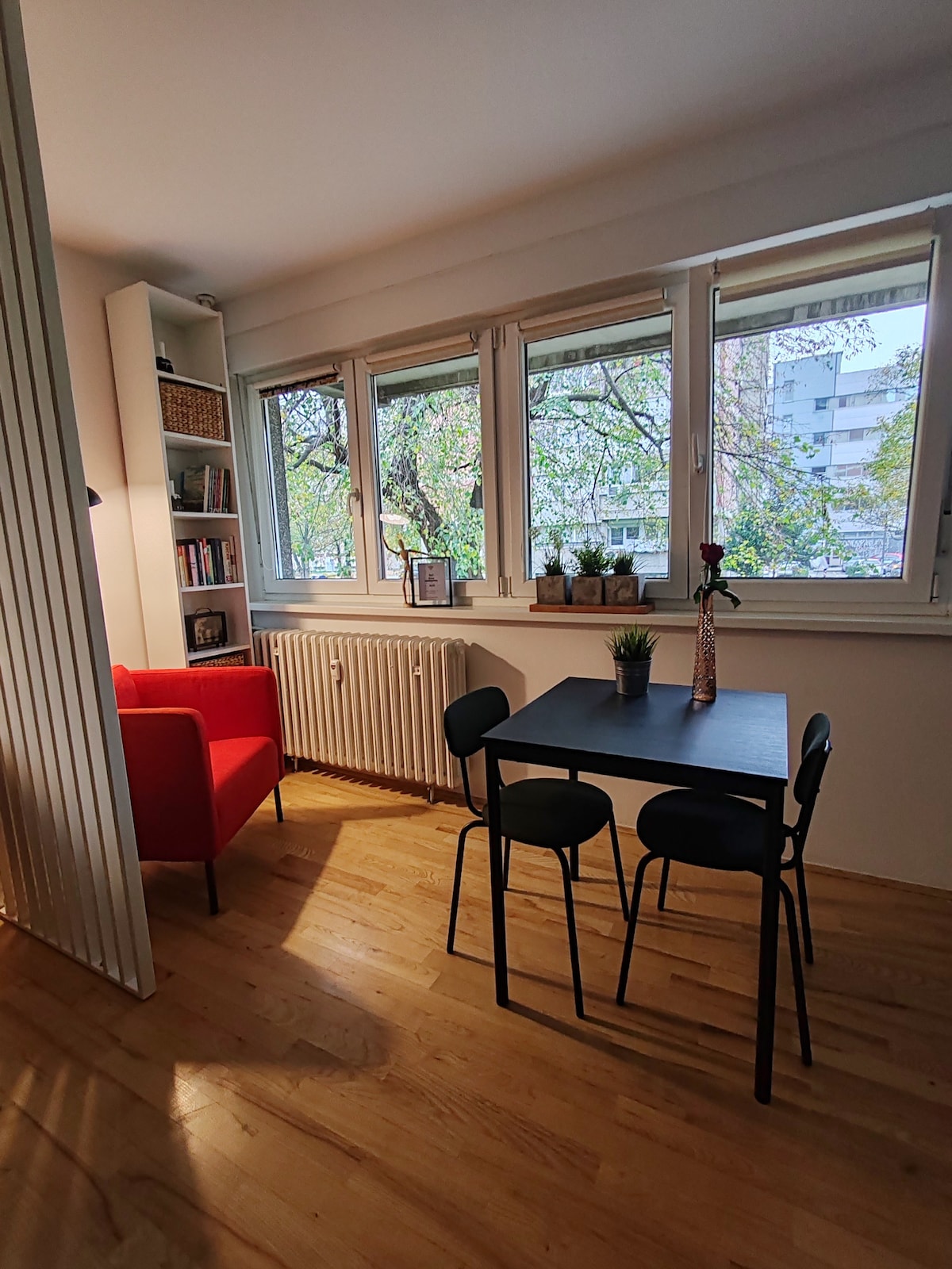A bright corner of the living area features large windows that provide natural light and a view of greenery outside. A small dining table with three black chairs is set within the space, complemented by a red armchair and a bookshelf on the side.