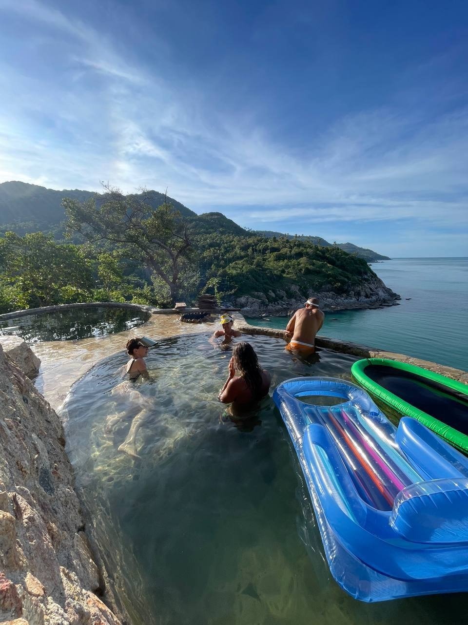 A stunning infinity pool overlooks the ocean and lush mountains. Several inflatable pool floats and individuals enjoying the serene surroundings are visible. The clear sky and calm waters create a peaceful ambiance, ideal for relaxation.