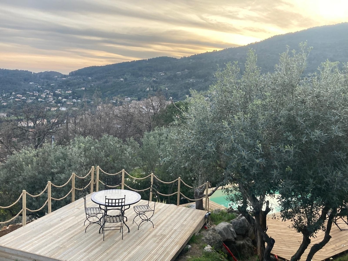 An outdoor terrace is revealed, featuring a circular table surrounded by four chairs. Olive trees dot the landscape, offering a natural setting. A distant view of the hills and valleys is visible under a soft sunset sky, adding depth to the serene environment.