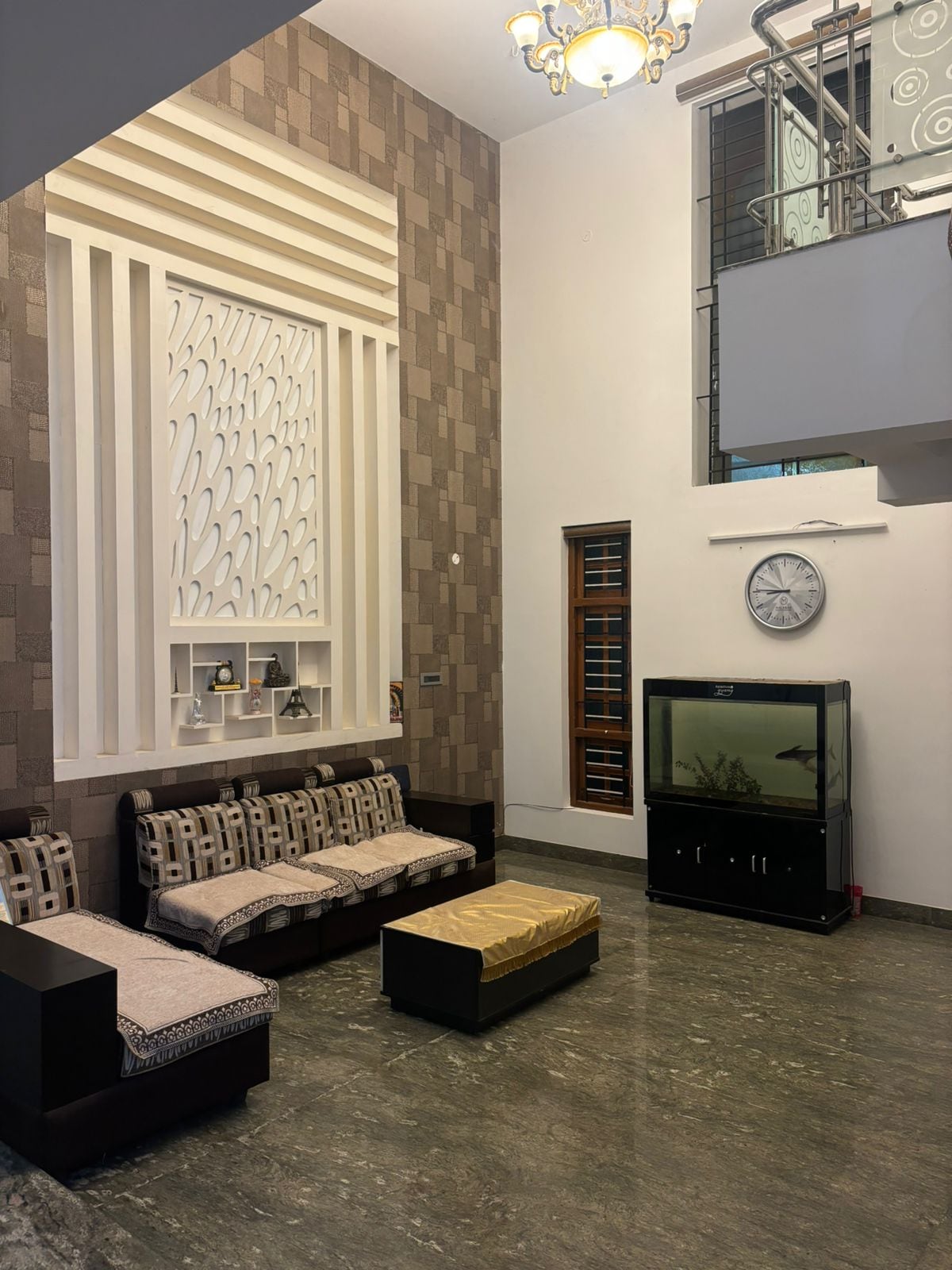 A spacious living area features a combination of comfortable seating and a television. Natural light floods the space through large windows, highlighting the minimalist design elements. A decorative wall with a unique pattern complements the modern decor, creating an inviting atmosphere.