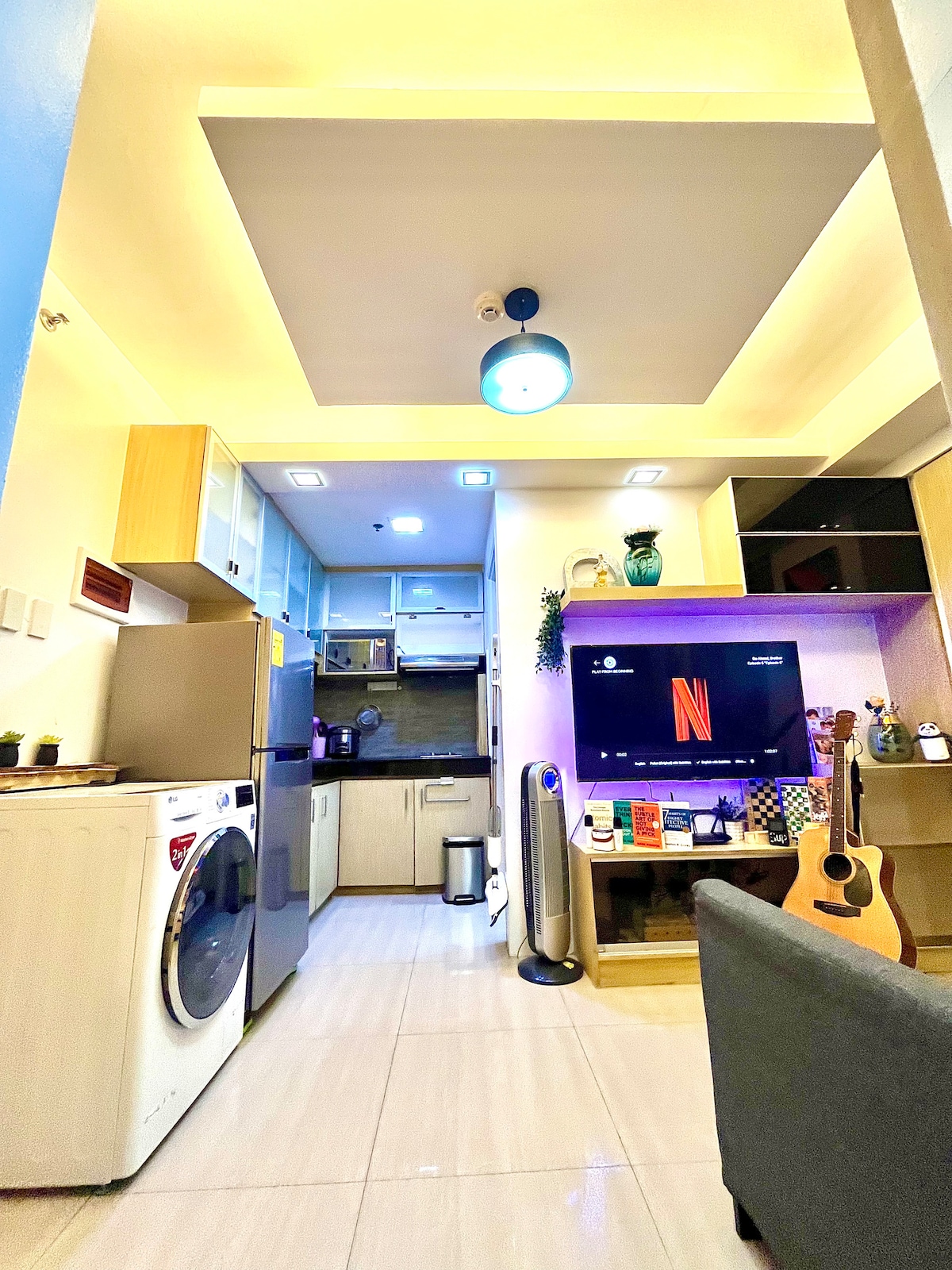 A modern kitchen space is showcased, featuring sleek cabinetry and a full-sized refrigerator. A washing machine is included for convenience. The living area is highlighted by a smart TV mounted on the wall, while a comfortable sofa adds to the inviting atmosphere.
