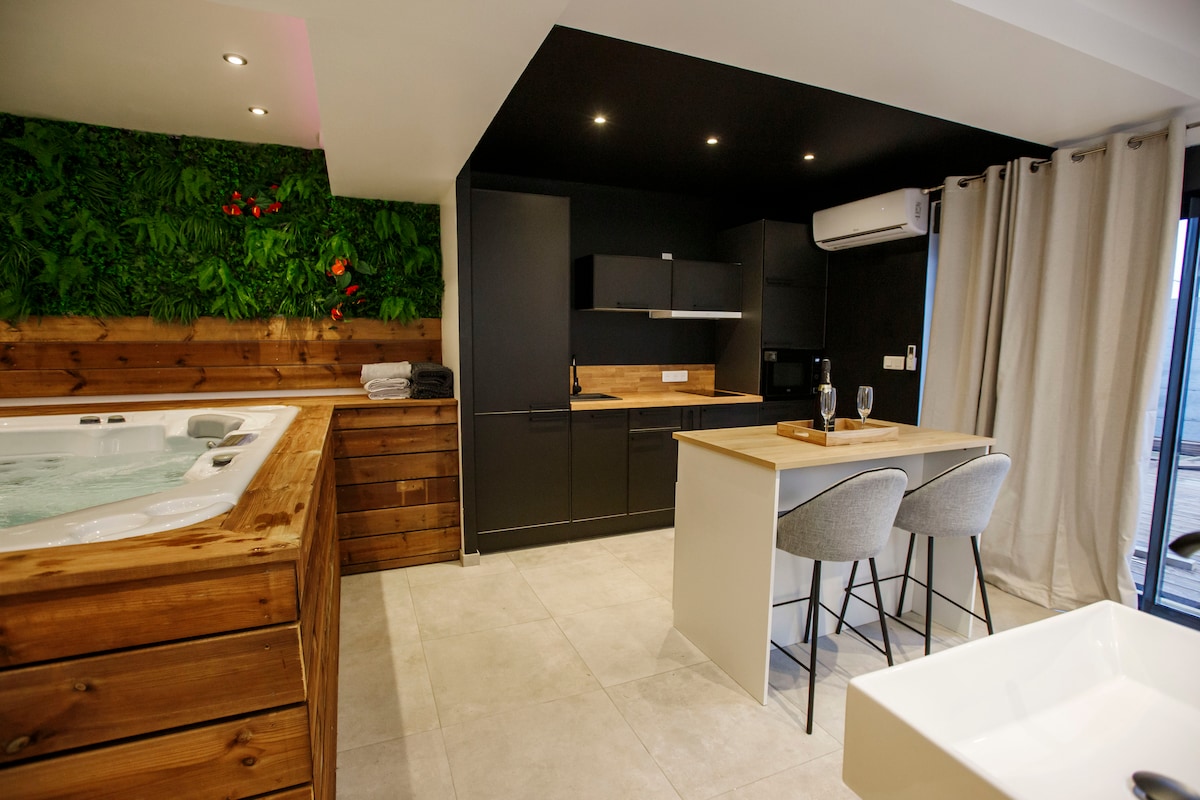 A modern kitchen is paired with a dining area featuring two stylish chairs and wooden countertops. A jacuzzi is positioned nearby, complemented by wooden cabinetry. The space is illuminated by soft overhead lights, and a wall adorned with greenery adds a touch of nature.