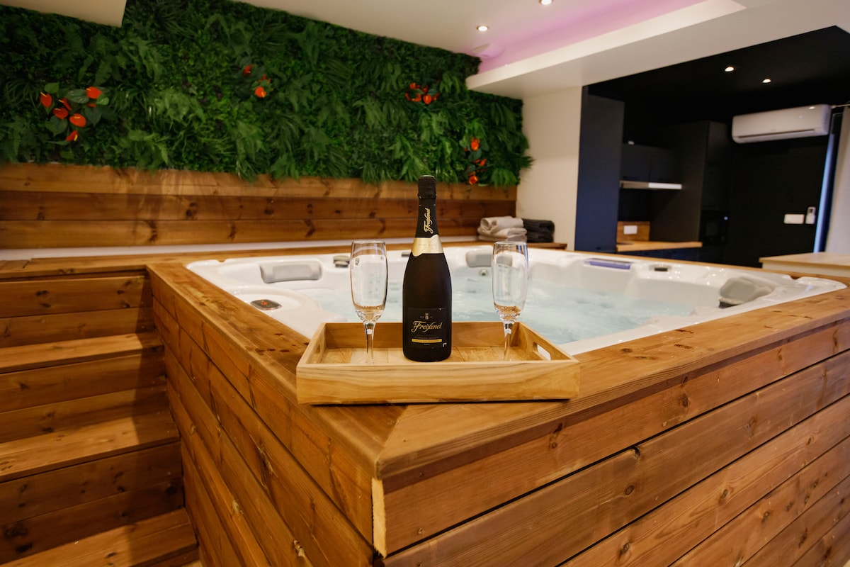 The jacuzzi serves as a focal point with wooden paneling and lush greenery in the background. A bottle of champagne and two glasses are placed on a wooden tray, inviting guests to enjoy a moment of relaxation. Soft lighting enhances the serene atmosphere.