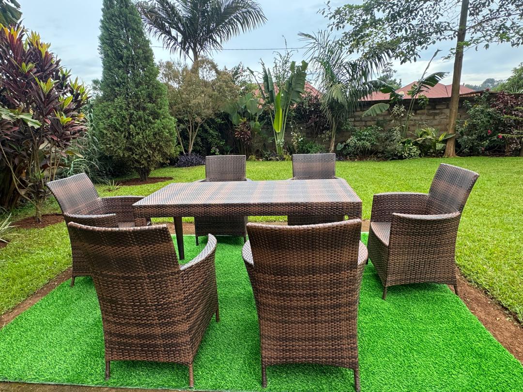 An outdoor seating area features a rectangular table surrounded by six wicker chairs. The space is set on a green grass mat, with tropical plants and trees providing a lush backdrop. The area is designed for relaxation and social gatherings.