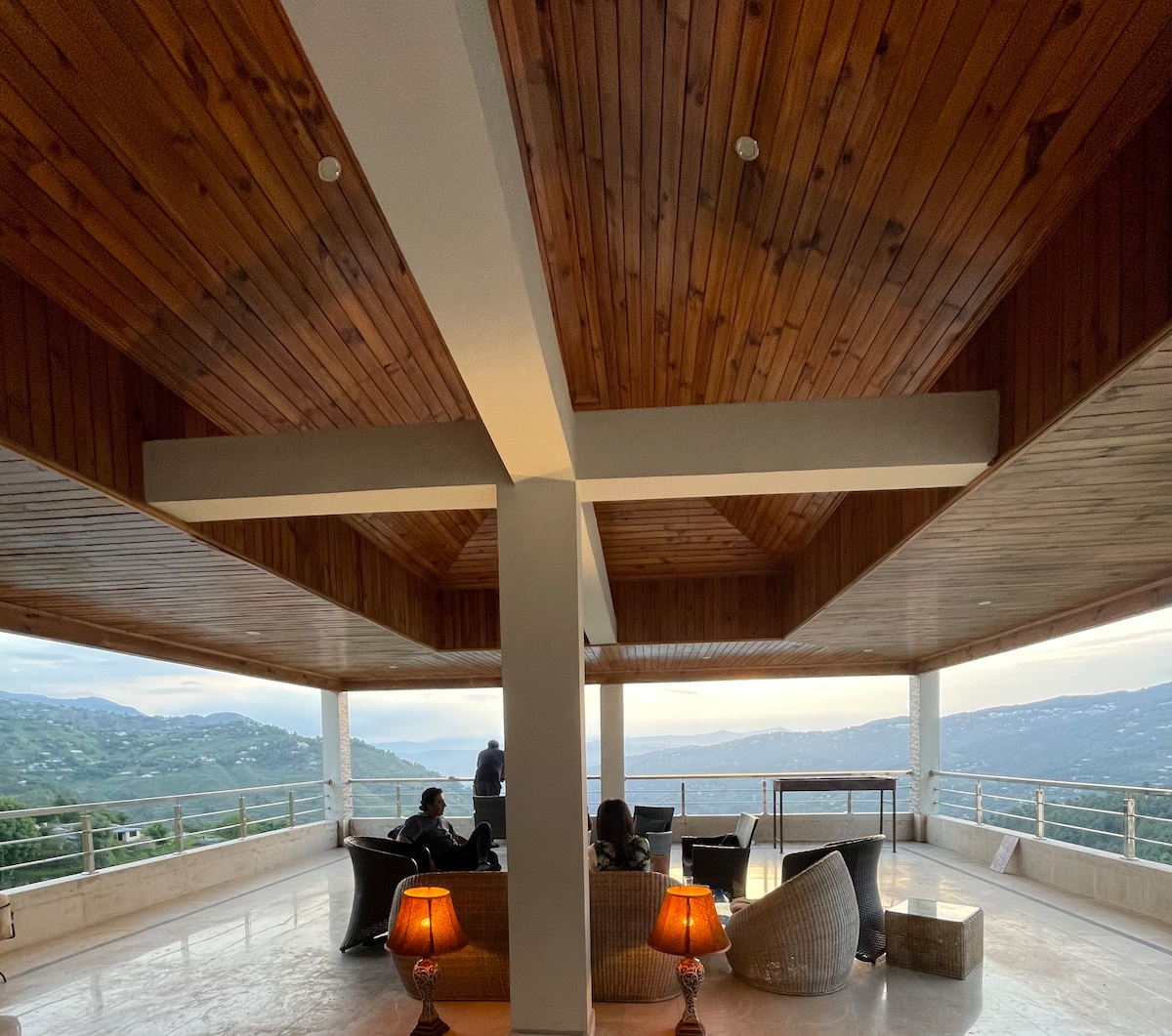 An expansive lounge area features a wooden ceiling with a crossbeam design, offering panoramic views of the hills. Comfortable seating arrangements are positioned for relaxation, complemented by two lamps providing ambient lighting. Large glass doors frame the surrounding landscape, enhancing the indoor-outdoor connection.