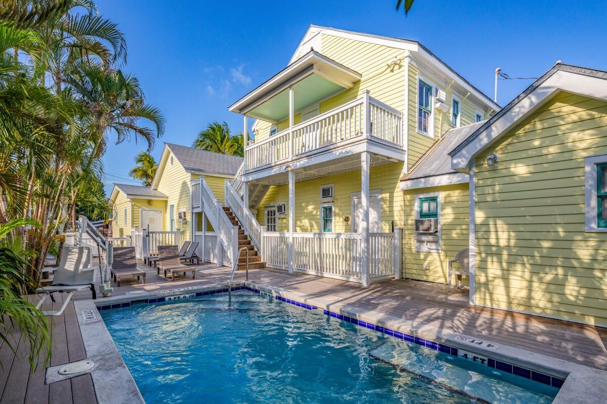 Bright yellow cottages are set against a clear blue sky, featuring a heated pool surrounded by wooden decking. Lush palm trees provide shade, while private balconies offer additional space for outdoor relaxation. Stairs lead up to the upper level, enhancing access to various outdoor areas.