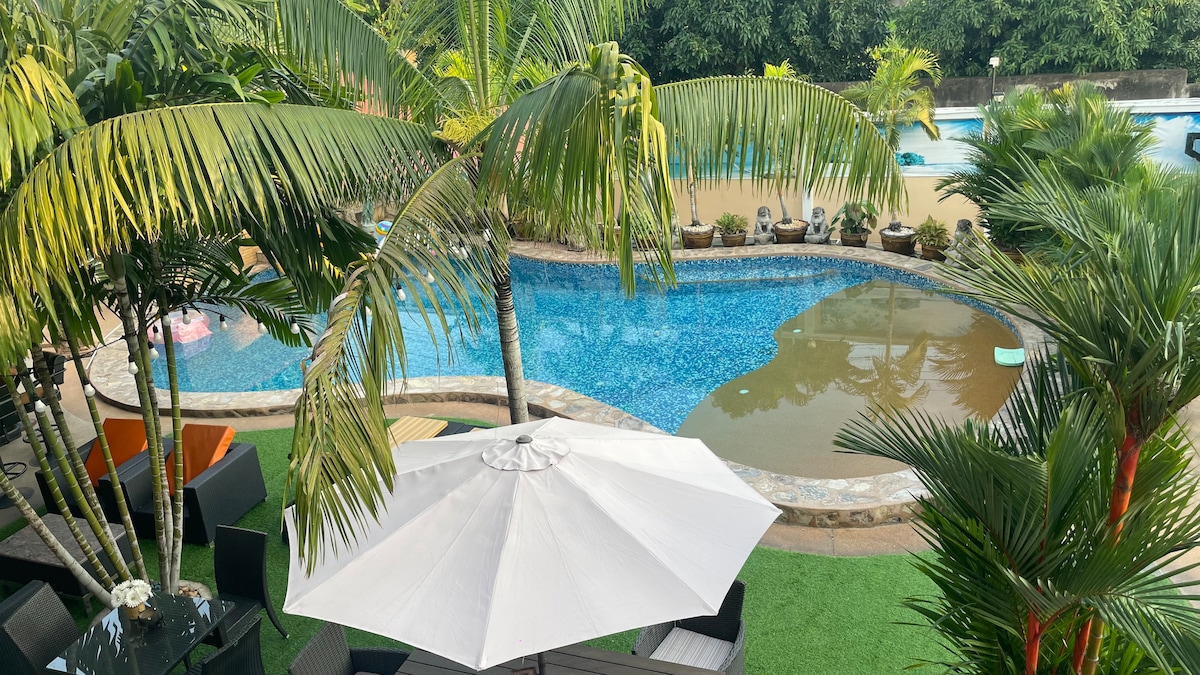 A spacious outdoor area features a shimmering swimming pool surrounded by lush palm trees. Comfortable lounge seating is arranged under a large white umbrella. The pool area includes gentle curves, enhancing the inviting atmosphere of this recreational space.
