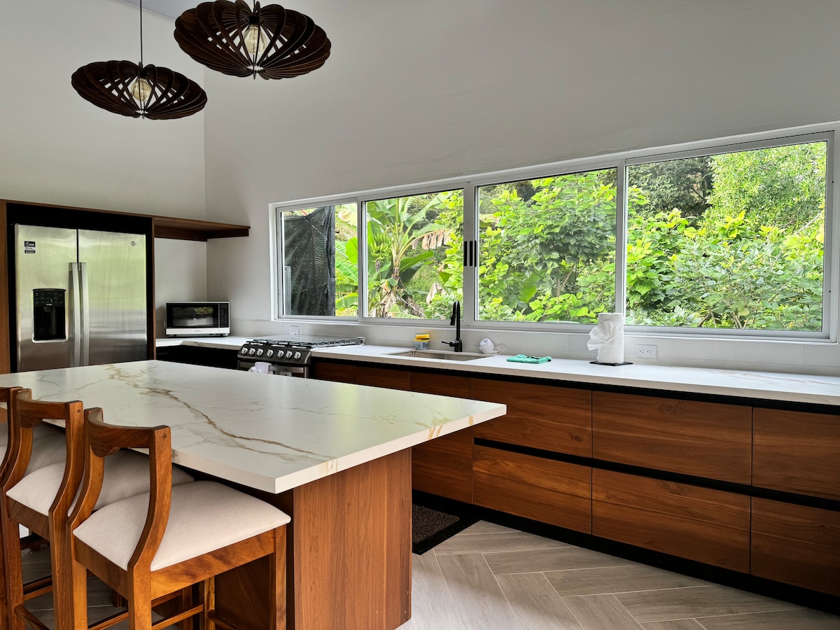 A modern kitchen area features a spacious island with a smooth countertop and wooden cabinets. Large windows provide a view of the surrounding greenery, while two distinct light fixtures hang above the island. Appliances include a refrigerator and a microwave, enhancing the functionality of the space.