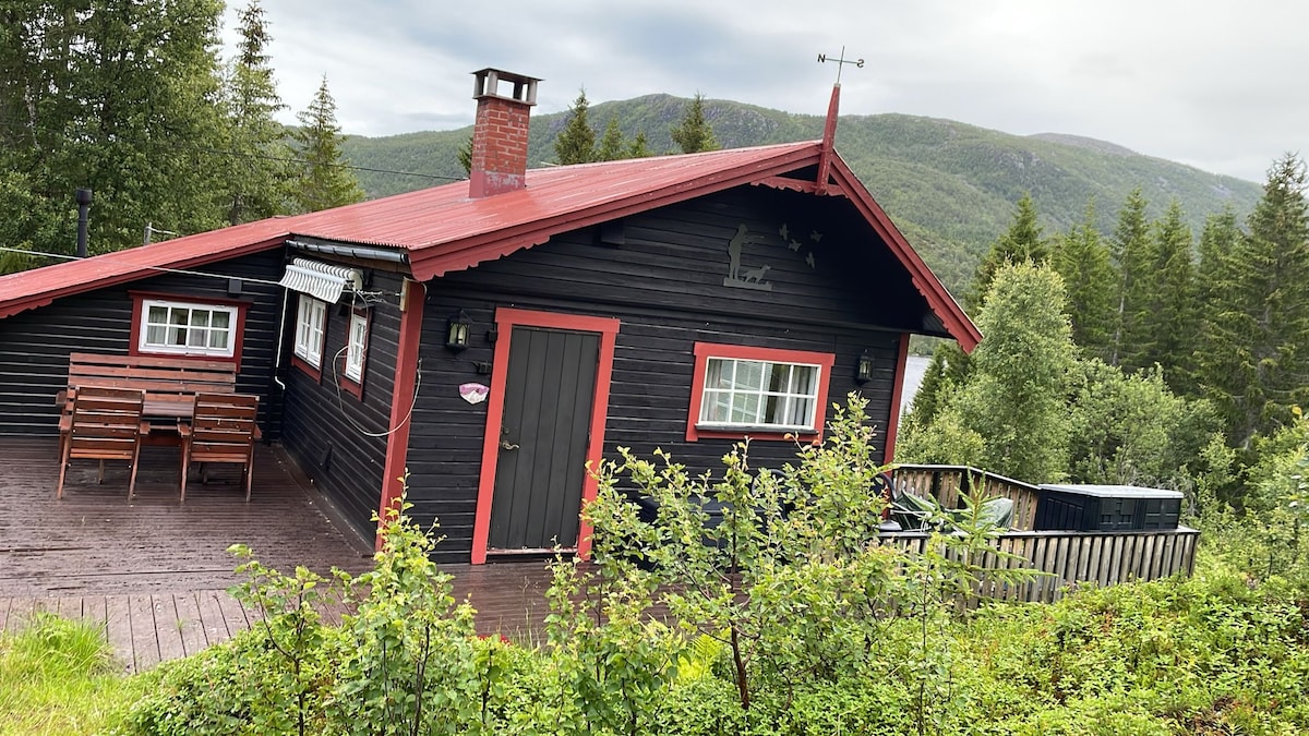 A cozy black wooden cabin with a red roof is surrounded by greenery. A deck with outdoor seating is visible, offering a scenic view of the nearby mountains and trees. A chimney adds a rustic touch to the cabin's exterior.