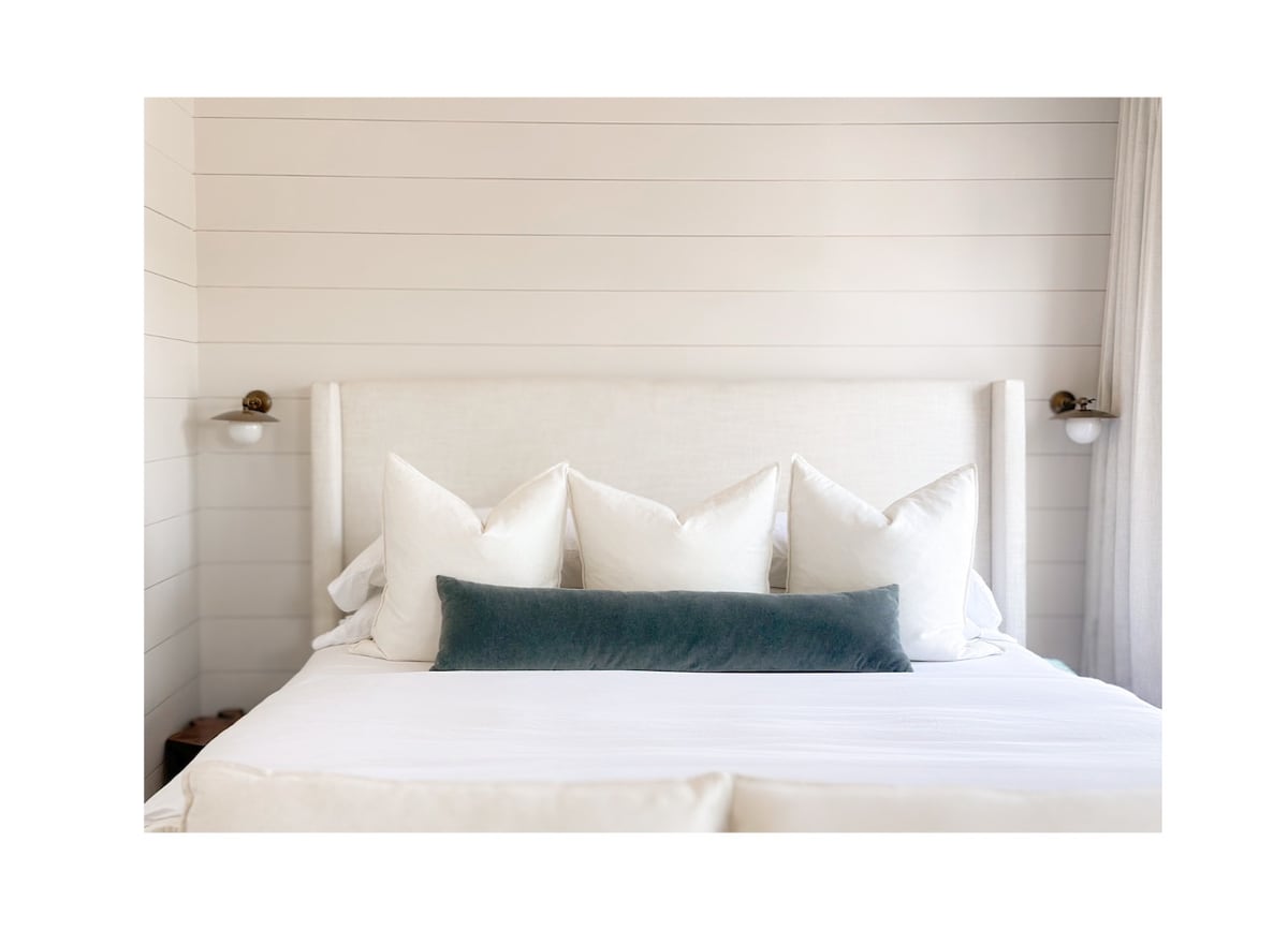 A spacious bed is positioned against a light wood-paneled wall, adorned with multiple white pillows showcasing varying textures. A long, soft teal pillow rests at the center, complementing the bedding. Two simple light fixtures are mounted on either side of the headboard.
