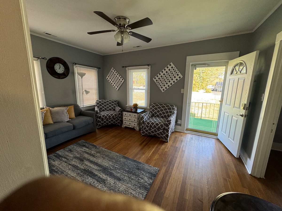 A living area is present, featuring a gray sofa and two patterned chairs arranged around a small, decorative table. Natural light enters through two windows, and a ceiling fan is mounted above. A door leads outside, providing access to a green space.