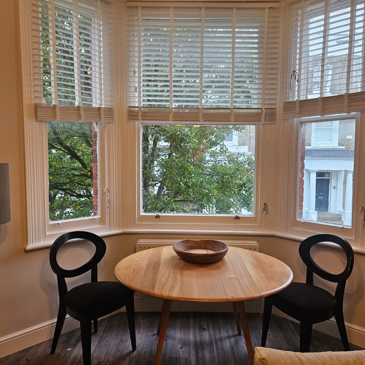 A small dining area is visible, featuring a wooden table set for two with a simple bowl in the center. Two black chairs frame the table, and light streams through the large bay windows adorned with white shutters, offering a view of greenery outside.