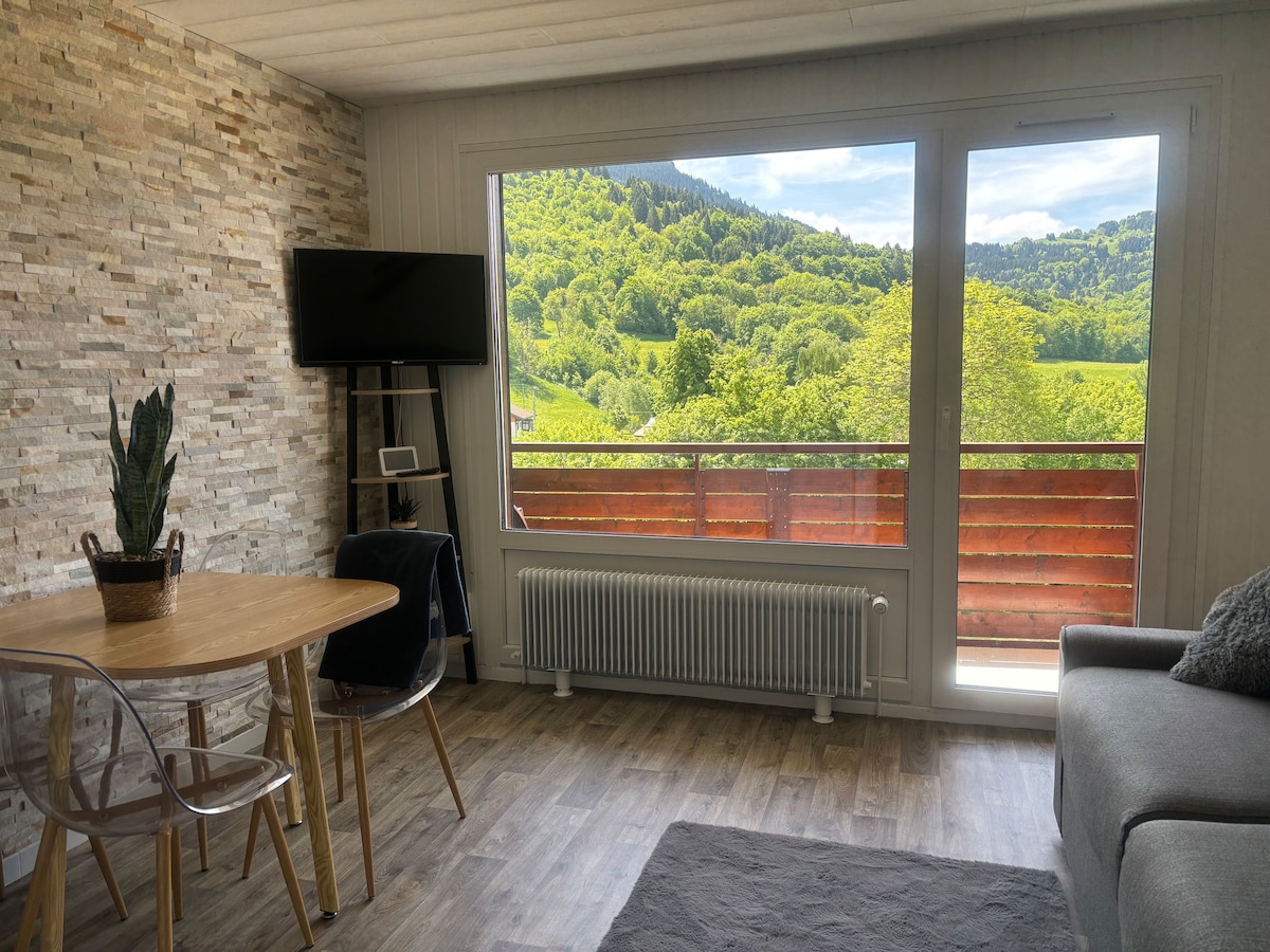 A modern living area is presented with a circular wooden dining table and transparent chairs. Large windows frame a scenic view of lush green hills. A flat-screen TV is mounted on the wall, and a cozy gray sofa is positioned nearby, creating an inviting space.