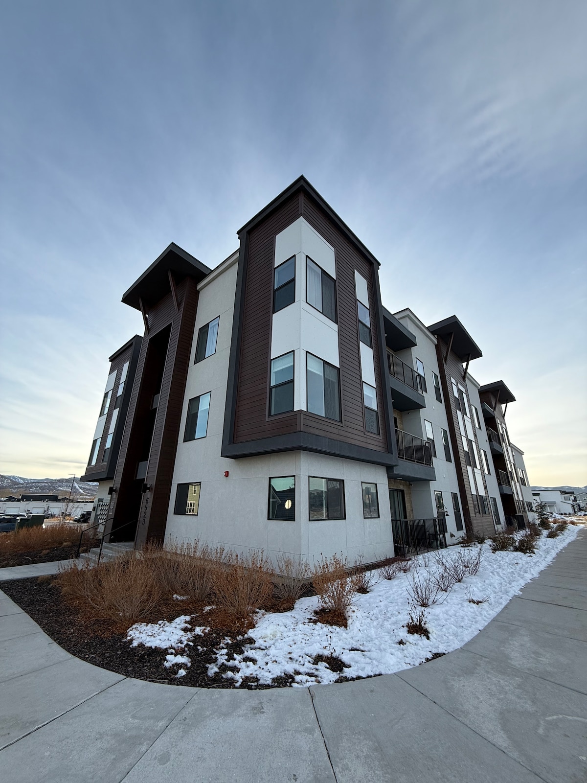 A modern three-story building features a combination of light and dark exterior finishes. Snow-covered landscaping lines the pathway leading to the entrance. Large windows provide a contemporary aesthetic while mountain views form a backdrop against a softly lit sky.